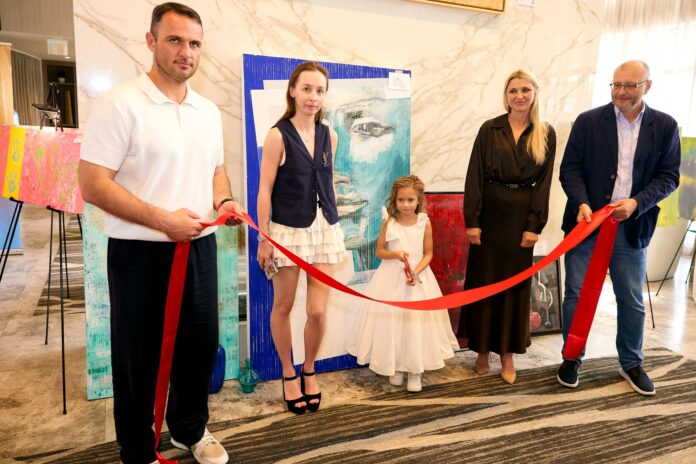 Five people stand in a lobby, cutting a red ribbon for a ceremony with colorful art panels in the background.