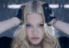 Madonna’s Confessions II Film Turns a New Album Rollout Into a Star-Studded Pop Event Close-up of a blonde woman with blue eyes, hands on her head, wearing fingerless gloves and a serious expression in moody lighting.