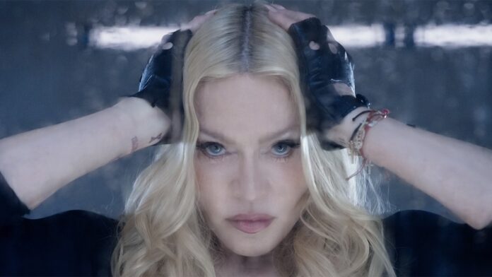 Close-up of a blonde woman with blue eyes, hands on her head, wearing fingerless gloves and a serious expression in moody lighting.