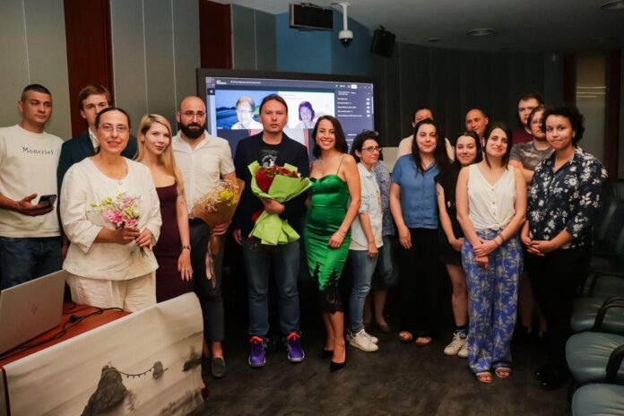 Group of people posing for a photo in a conference room, some holding bouquets, with a presentation screen in the background.