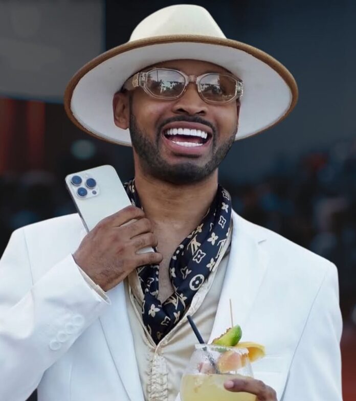 Man in a white suit and hat, smiling, holding a smartphone and a citrus cocktail.