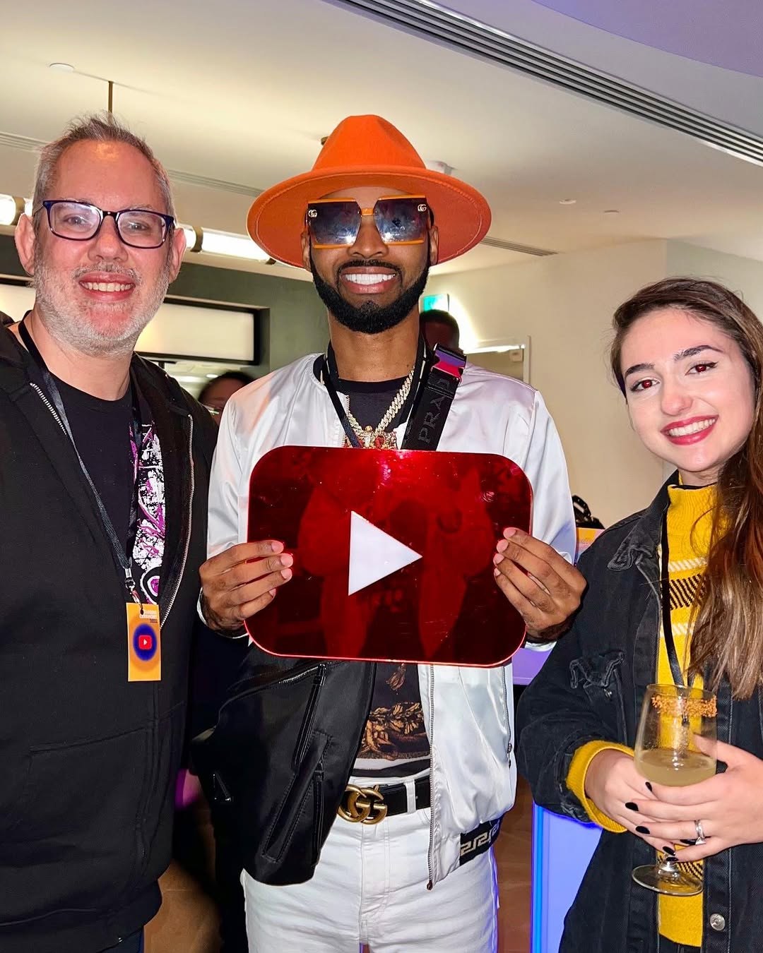 Three people posing together; the man in the center holds a red YouTube Play button plaque and wears an orange hat and white outfit, flanked by two friends in a lively setting.
