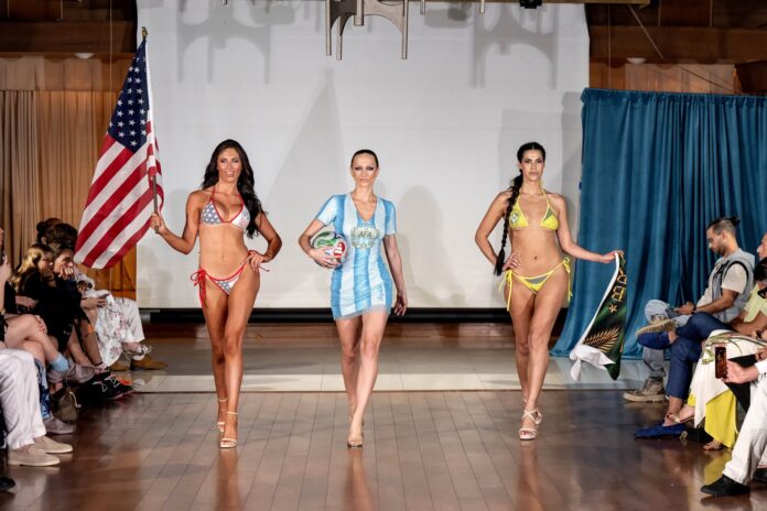 Three models walk a runway during a beachwear show: left model holds an American flag in a red-white bikini; center model in blue-and-white striped dress holds a volleyball; right model in yellow bikini poses with a green sarong as spectators watch.